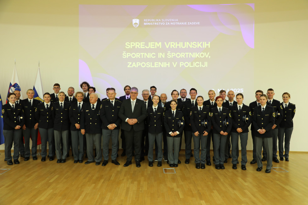 Okrog 50 ljudi stoji v treh vrstah. Večina je v policijskih uniformah. Za njimi je na steni projekcija.